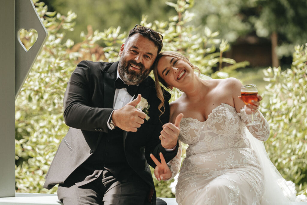 Amidst lush greenery, a bride and groom share joyful smiles and thumbs-up. The groom dons a black suit with a bow tie, while the bride dazzles in an off-shoulder lace wedding dress, holding a drink. Captured by skilled wedding photography, their happiness is both vibrant and timeless. © Aimee Lince Photography