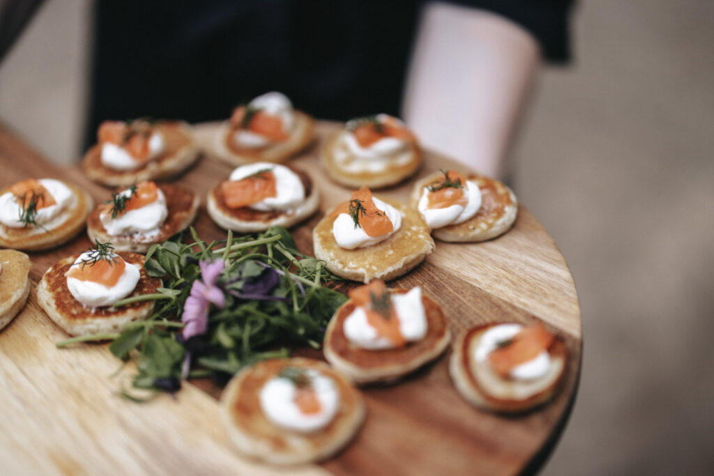 A wooden platter holds several bite-sized appetizers, perfect for a wedding photographer's lens. Each consists of a small pancake topped with cream, smoked salmon, and dill. In the center is a garnish of greens and a purple flower. A person's arm is seen in the background, holding the platter at Elsham Hall. © Aimee Lince Photography