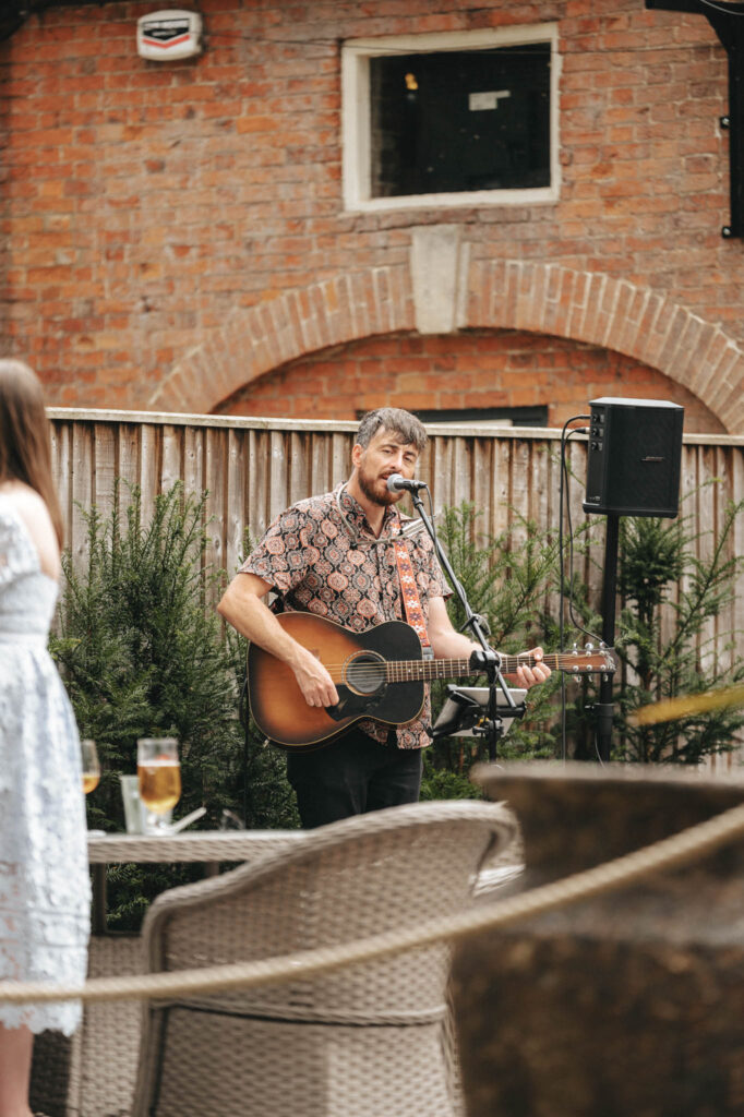 A man with a gray beard strums an acoustic guitar, singing into a microphone outdoors at Elsham Hall. He's in front of a wooden fence, donning a patterned shirt. Nearby, a woman in a white dress stands with drinks on the table, embodying the perfect scene for any wedding photographer. © Aimee Lince Photography