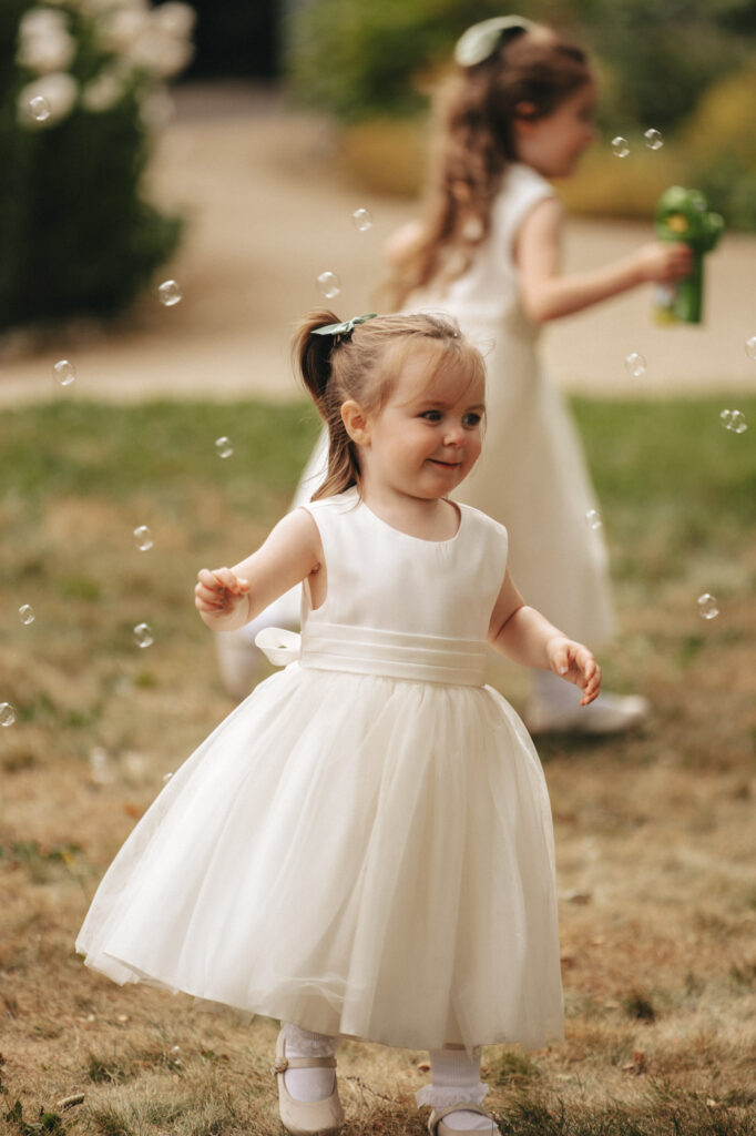 At Elsham Hall, a young girl in a white dress joyfully explores the garden, surrounded by bubbles captured through exquisite photography. Her hair is tied with a bow as she smiles. In the background, another child in a similar dress wields a bubble blower amidst lush greenery and a charming path. © Aimee Lince Photography