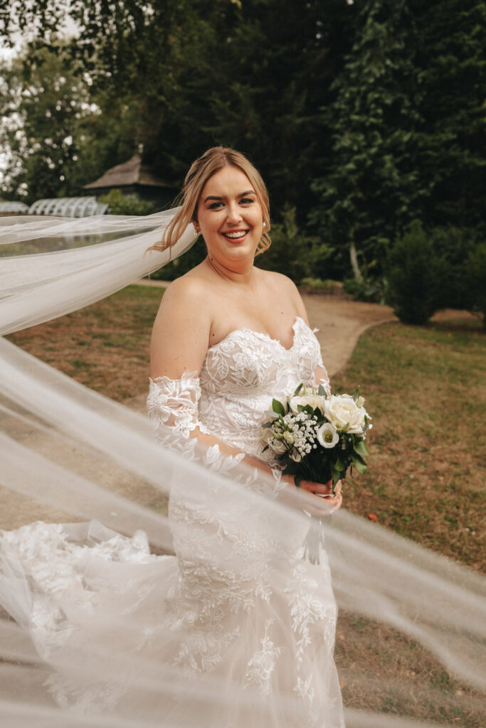 A bride in an off-the-shoulder lace wedding dress smiles while holding a bouquet of white flowers and greenery. Her veil flows in the breeze as she stands on a grass path at Elsham Hall, with lush greenery and a rustic building in the background, captured by her talented wedding photographer. © Aimee Lince Photography