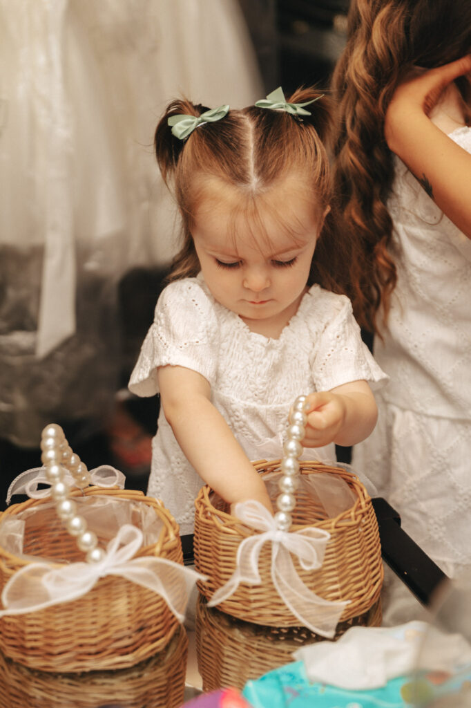 A young girl with pigtails and green bows is dressed in a white lace dress, captured beautifully by the photographer. She gently places a pearl necklace into a woven basket adorned with a white ribbon. Another child with long hair is partially visible beside her, also wearing a white dress. © Aimee Lince Photography