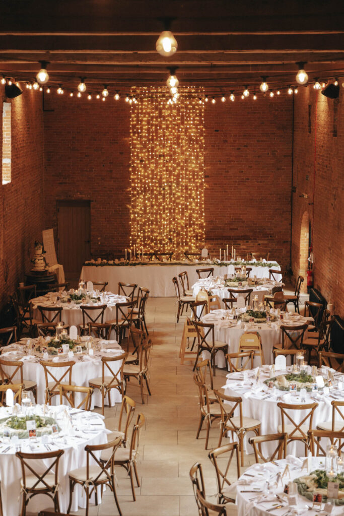 Wedding reception in a rustic brick hall at Elsham Hall with round tables set for guests. Tables are draped in white tablecloths, adorned with greenery and candles. Warm string lights hang from the wooden ceiling beams, creating a cozy atmosphere perfect for photography. A backdrop of fairy lights decorates one end of the room. © Aimee Lince Photography