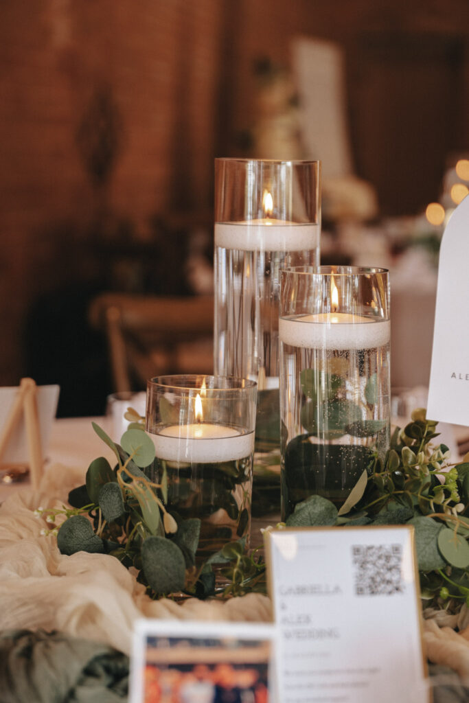 Tall glass vases with floating candles sit on a table amidst lush greenery, capturing the eye of any discerning wedding photographer. The varying heights and warm glow create a picturesque scene, enhanced by a white card featuring text and QR codes. The elegant setting suggests a formal event like a wedding. © Aimee Lince Photography