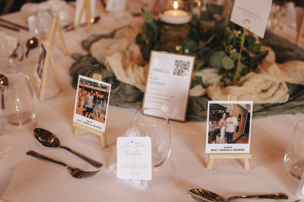 A wedding table setting showcases small easels with Polaroid photos—a nod to timeless photography—alongside a menu card. Votive candles, lush greenery, a glass with a place card, and elegant tableware create an inviting ambiance perfect for capturing by any wedding photographer. © Aimee Lince Photography