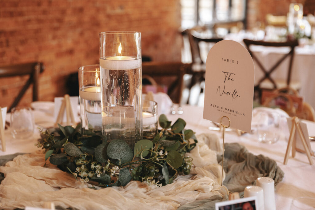 At Elsham Hall, a wedding table centerpiece captures the eye with a trio of floating candles in tall glass vases, surrounded by eucalyptus leaves and delicate white flowers. A card reads, “Table 2, The Veranda, Alex & Gabriella.” The scene is perfect for photography against the brick wall backdrop. © Aimee Lince Photography