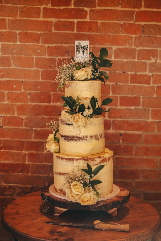 A three-tiered semi-naked cake, elegantly adorned with white roses, greenery, and baby's breath, rests on a round wooden base. A small photo topper crowns it. This wedding masterpiece is beautifully captured by the photographer against a rustic brick wall backdrop. © Aimee Lince Photography