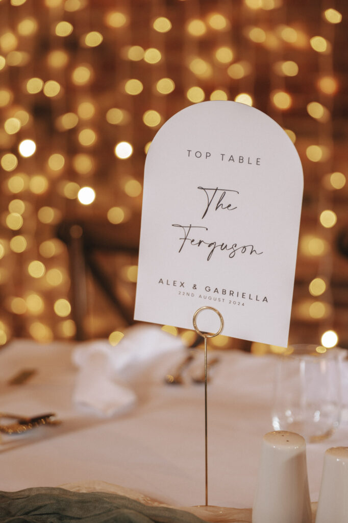 Wedding table card with "Top Table" and "The Ferguson" in elegant script, set against a backdrop of warm, golden fairy lights. Below, "Alex & Gabriella" and "22nd August 2024" are printed. Captured beautifully by a wedding photographer at Elsham Hall, it rests on a stand with blurred tableware in the foreground. © Aimee Lince Photography