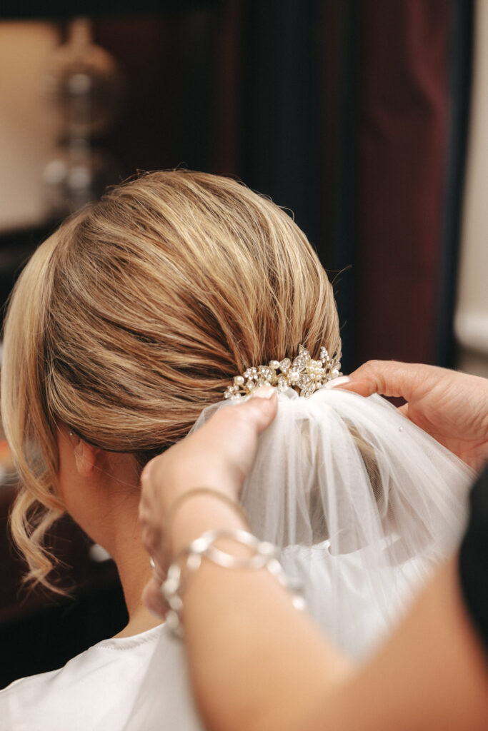 A bride's hair is styled in an elegant bun adorned with a sparkling hairpiece, while a photographer captures someone attaching a white tulle veil. The intricate details of the hairstyle and accessories shine through against the softly blurred background, enhancing this beautiful wedding moment. © Aimee Lince Photography