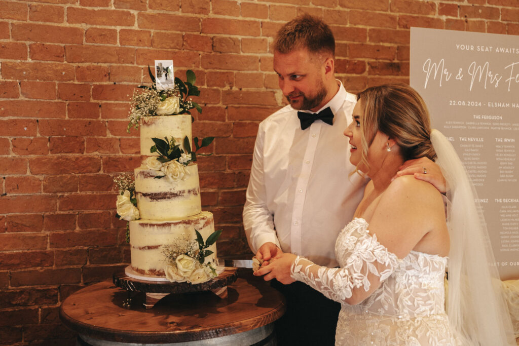 A bride and groom stand together at Elsham Hall, cutting a tall, rustic three-tiered naked cake adorned with white roses and greenery. The bride wears an off-the-shoulder lace gown, and the groom sports a white shirt with a black bow tie. A wedding photographer captures this moment in the brick-walled room with a seating chart visible. © Aimee Lince Photography