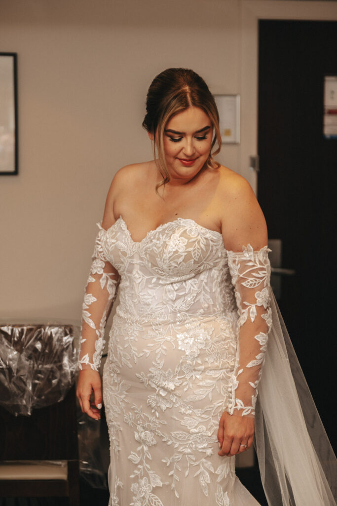 Bridal portrait of a woman in a white, off-the-shoulder lace gown with floral embroidery and sheer long sleeves by a talented wedding photographer. Her hair is styled in an updo. She smiles softly, standing indoors with a dark door and framed picture at Elsham Hall in the background. © Aimee Lince Photography