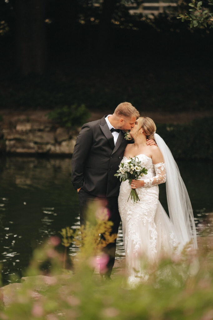 A bride in a lace gown and veil holds a bouquet, kissing a groom in a black suit with a bow tie. Captured by a wedding photographer at Elsham Hall in Grimsby, they stand by a calm pond with greenery. Surrounded by soft, blurred flowers, the romantic scene evokes an intimate and serene atmosphere. © Aimee Lince Photography