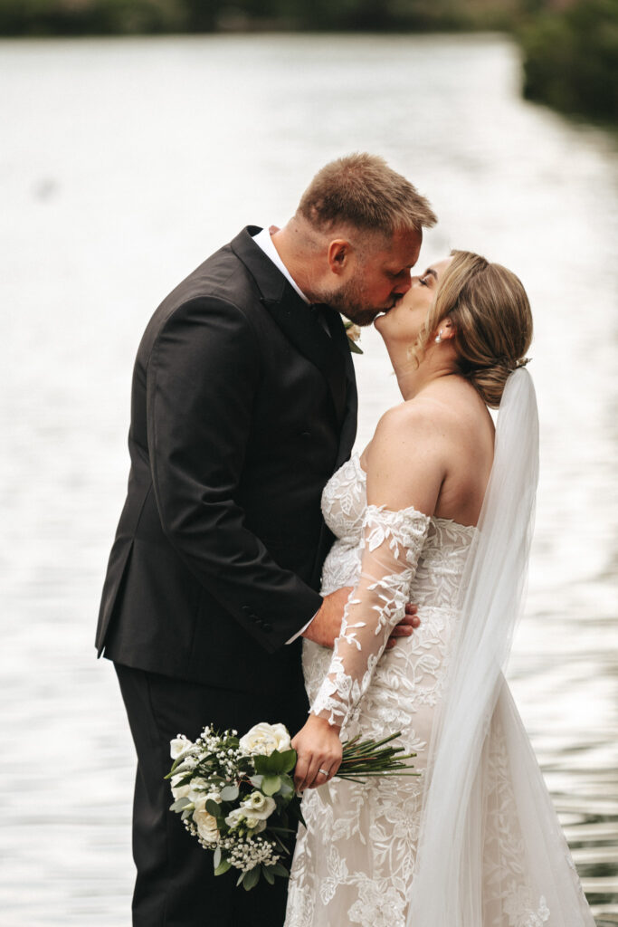 A couple kisses by a serene lakeside at Elsham Hall. The bride, in her white lace off-shoulder gown, holds a bouquet of white flowers, while the groom dons a black suit. Their photographer captures the tranquil and romantic ambiance set by the trees and water, crafting a timeless memory. © Aimee Lince Photography