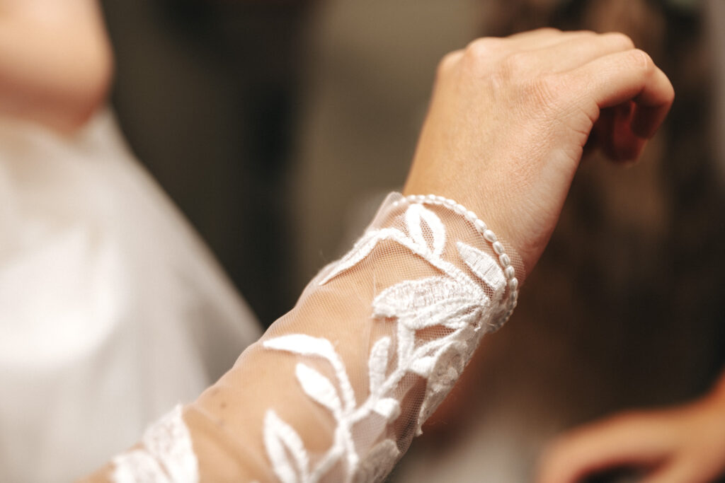 A close-up of a person's hand, raised. They wear a long-sleeved garment with sheer fabric featuring white embroidered floral designs. The sleeve is delicately beaded at the wrist, suggesting formal or bridal attire. Captured by a skilled photographer, the background is softly blurred. © Aimee Lince Photography