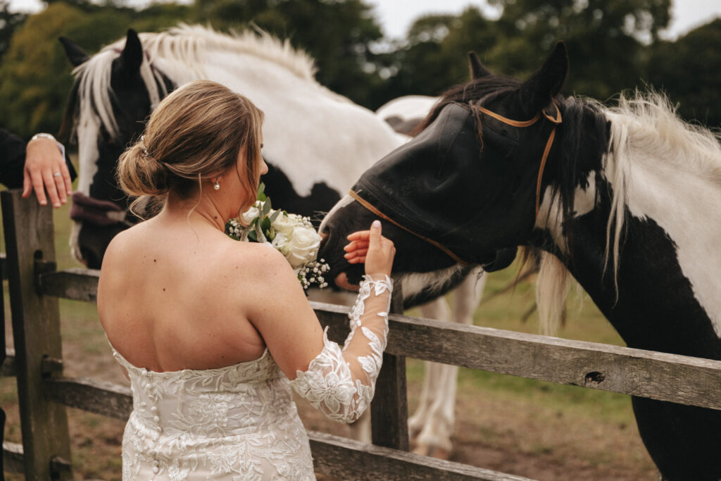 A bride in a white lace wedding dress with off-the-shoulder sleeves stands near a wooden fence at Elsham Hall. She feeds a bouquet of white flowers to a black and white horse wearing a fly mask, as another horse waits nearby, all beautifully captured by the wedding photographer against the verdant backdrop. © Aimee Lince Photography