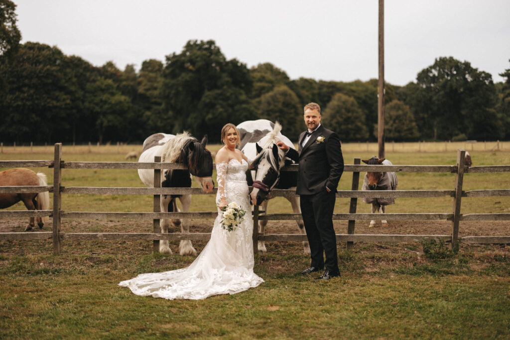 A bride in a lace wedding dress and a groom in a black suit stand smiling near a wooden fence, captured beautifully by the photographer. The bride holds a bouquet while behind them, a black and white horse and brown pony grace the grassy field with trees under an overcast sky—a perfect wedding moment. © Aimee Lince Photography