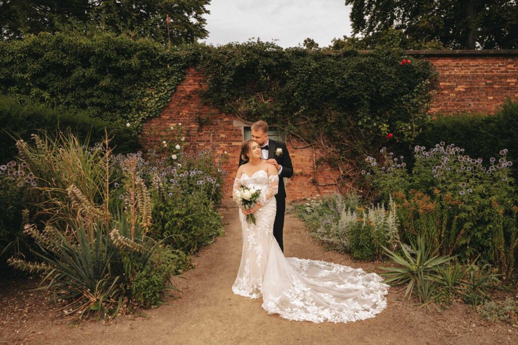 Bride in a white, off-the-shoulder gown with a long lace train and groom in a black tuxedo stand in the lush gardens of Elsham Hall. He's gently embracing her from behind. Surrounded by green plants, flowers, and an ivy-covered brick wall, the scene is perfect for wedding photography. © Aimee Lince Photography