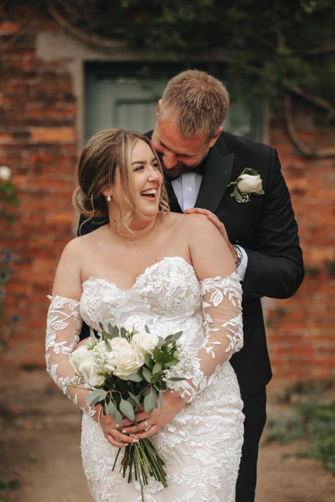 A bride in a white lace dress holds a bouquet of white roses, smiling joyfully for the wedding photographer. Her groom, in a black suit with a white rose boutonniere, stands behind her, affectionately leaning over her shoulder. They pose outdoors at Elsham Hall in Lincolnshire with a brick wall and greenery in the background. © Aimee Lince Photography