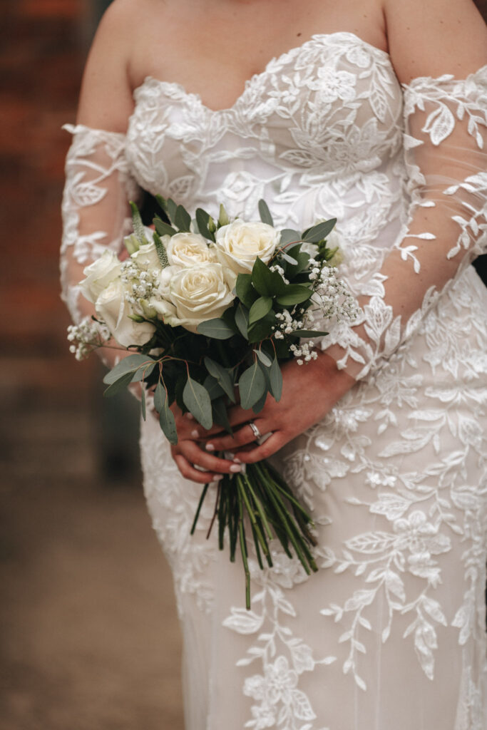 In a softly blurred background, a bride in an off-the-shoulder, lace wedding gown captures the essence of the day. Holding a bouquet of white roses adorned with green leaves and baby's breath, her look is beautifully framed by the keen eye of an expert wedding photographer. © Aimee Lince Photography
