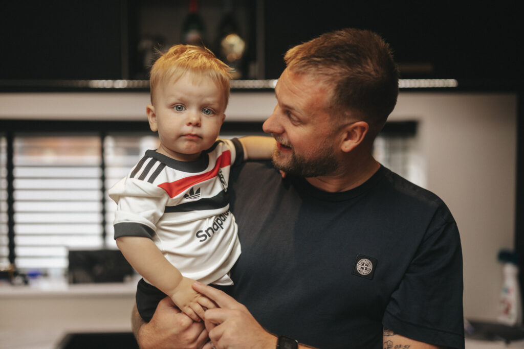 A man with short hair, wearing a dark t-shirt, holds a toddler dressed in a soccer jersey. The toddler looks at the camera while the man smiles at him. They are indoors with a blurred, photographically composed background of a modern kitchen and bottles on a shelf. © Aimee Lince Photography