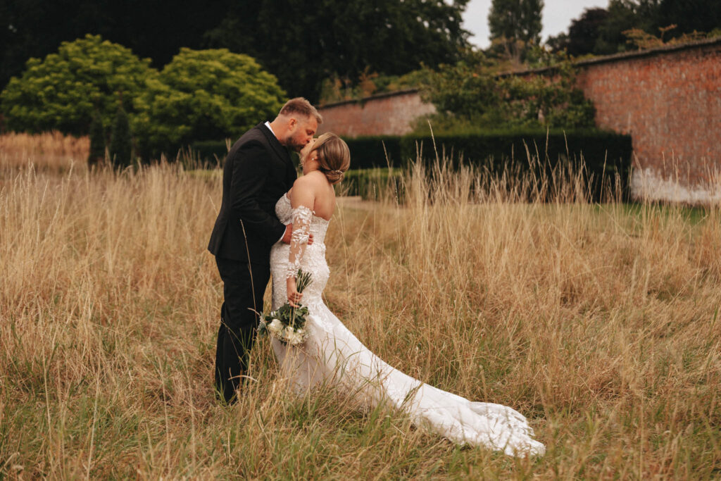 A bride and groom share a kiss in a grassy field, beautifully captured by a wedding photographer. The bride's white, off-the-shoulder gown with lace details complements her bouquet, while the groom stands elegantly in his black suit. A red brick wall and green trees form the serene, romantic backdrop. © Aimee Lince Photography