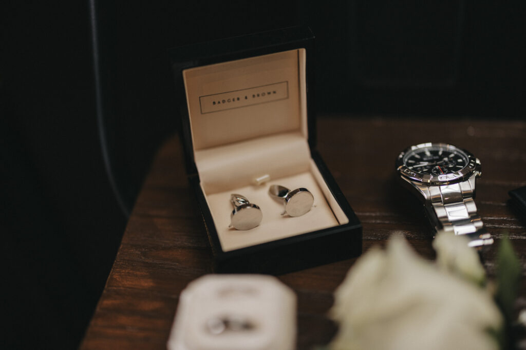A pair of silver cufflinks in an open black box with a white interior marked "Badger & Brown" sits elegantly on a wooden surface, captured with the precision only a skilled photographer can achieve. Beside it is a silver wristwatch and a delicate white flower, with another small white box visible in the foreground. © Aimee Lince Photography