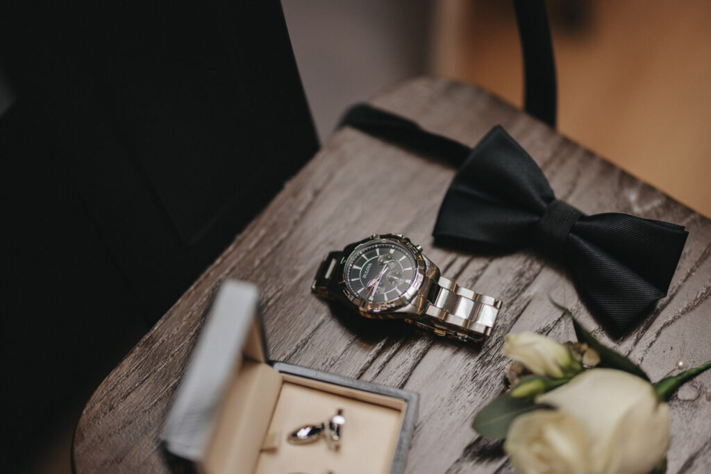 A polished silver watch with a black face is placed on a wooden table beside a black bow tie, captured perfectly by the wedding photographer. Nearby, a pair of cufflinks rest in an open box, and a white rose boutonniere lies on the table, creating an elegant and formal arrangement. © Aimee Lince Photography