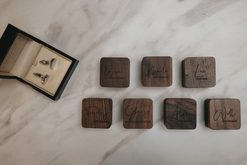 A set of cufflinks in an open black box rests on a marble surface, capturing the rustic, elegant feel perfect for wedding photography. Nearby, seven wooden coasters engraved with names and titles like "Groomsman" and "Best Man" add charm to this memorable scene at Elsham Hall. © Aimee Lince Photography