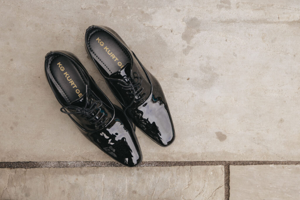 A pair of glossy black leather dress shoes with pointed toes and laces is artfully captured by a photographer on a textured stone surface, perfect for a wedding. The brand name "Kurt Geiger" graces the insoles, adding an elegant touch reminiscent of Elsham Hall's timeless charm. © Aimee Lince Photography