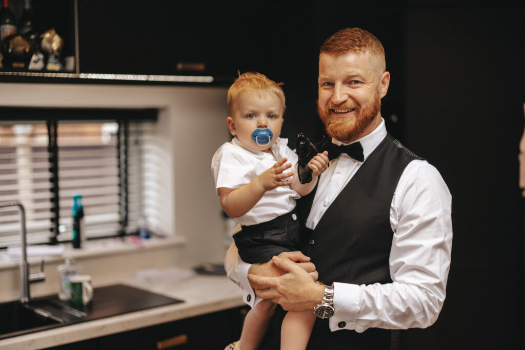 A bearded man, resembling a wedding photographer in his formal outfit with a bow tie and vest, holds a toddler in a kitchen. The toddler wears shorts and a white shirt, clutching a toy and pacifier. Black cabinets line the room as natural light streams through blinds onto the countertop. © Aimee Lince Photography