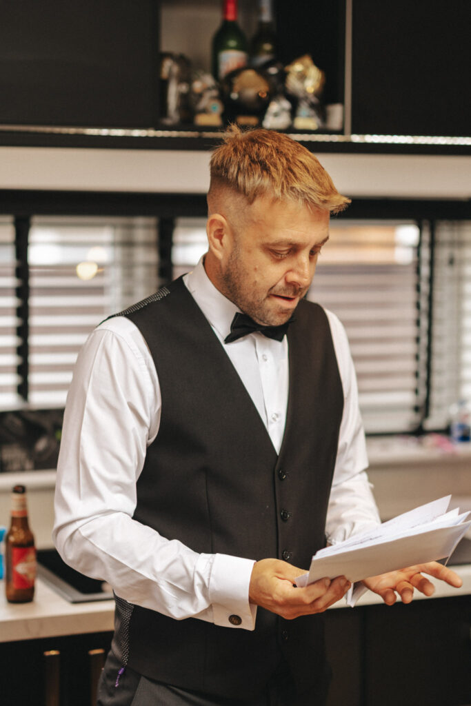 A man with short hair, wearing a black vest, white shirt, and bow tie, stands indoors reading papers. Looking as if he's preparing for a wedding shoot, maybe even practicing photography poses, he seems focused. Behind him is a counter with bottles and decor; blinds cover the windows in the background. © Aimee Lince Photography