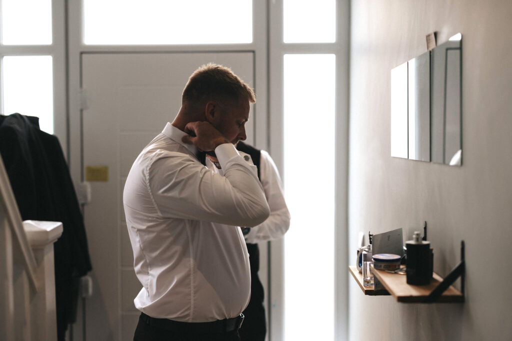 A man in a white shirt, possibly a wedding photographer, adjusts his tie in a hallway with a wall-mounted mirror. Another person, slightly blurred, stands in the background at Elsham Hall. Coats hang on a banister, and assorted items are scattered on a shelf. Soft light filters through nearby windows. © Aimee Lince Photography