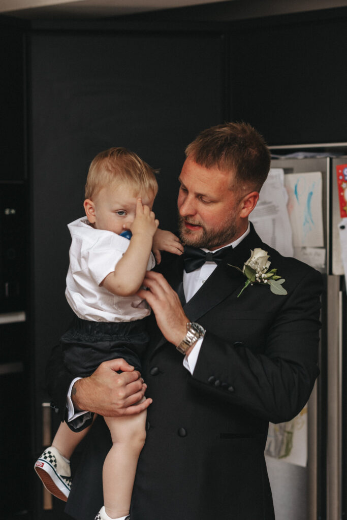 A man in a black suit with a boutonniere holds a young child, likely captured during wedding photography. The child, dressed in a white shirt and black shorts, covers part of their face with their hand. They stand in a room decorated with drawings and magnets on the refrigerator. © Aimee Lince Photography