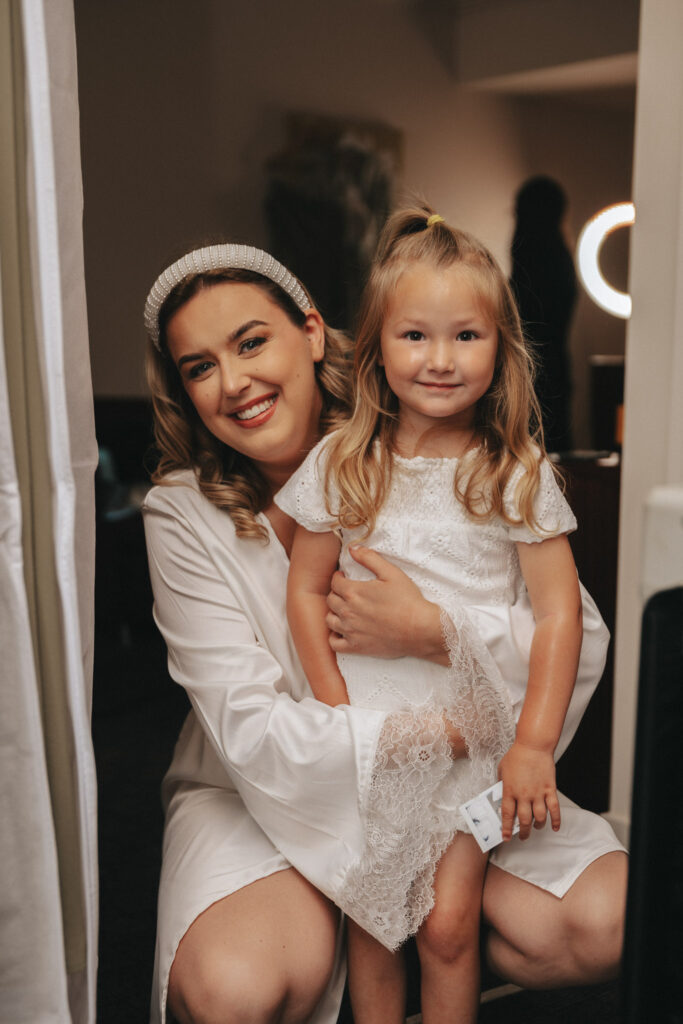 A woman smiling and kneeling next to a young girl in a white lace dress. The woman, perhaps a wedding photographer, wears a headband and a white robe. In the softly lit room of Elsham Hall, a mirror with round lights shines in the background, and the girl has a small bow in her hair. © Aimee Lince Photography