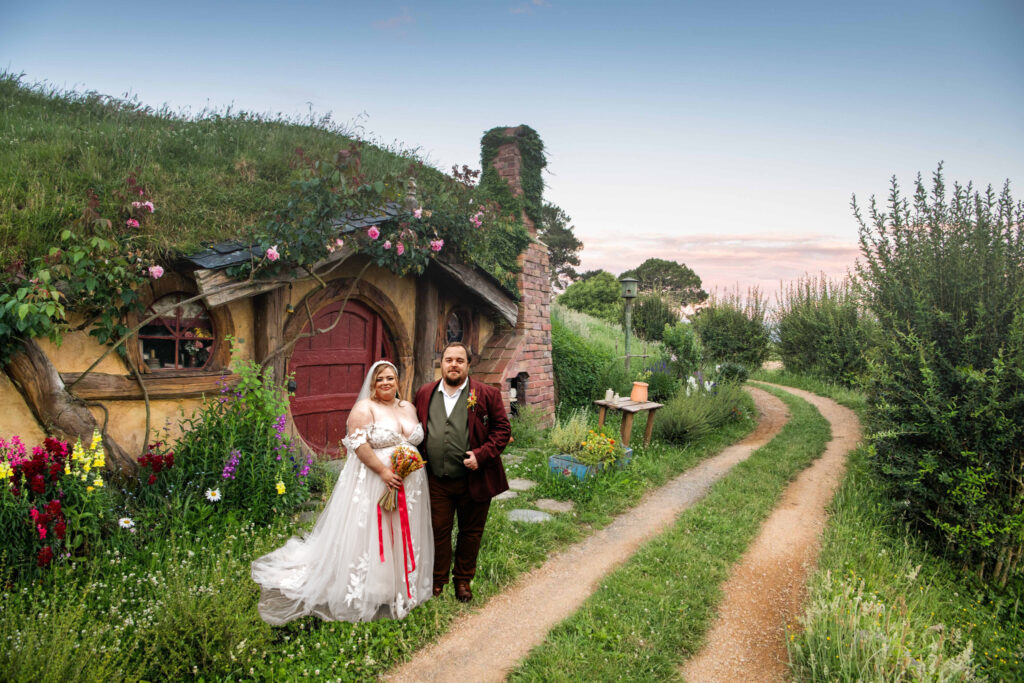 A bride and groom stand outside a hobbit-like house with a red door, reminiscent of The Shire from Lord of the Rings, surrounded by lush greenery and vibrant flowers. The bride wears a white gown and holds a bouquet, while the groom is in a brown suit. A winding path leads away under a clear sky. © Aimee Lince Photography