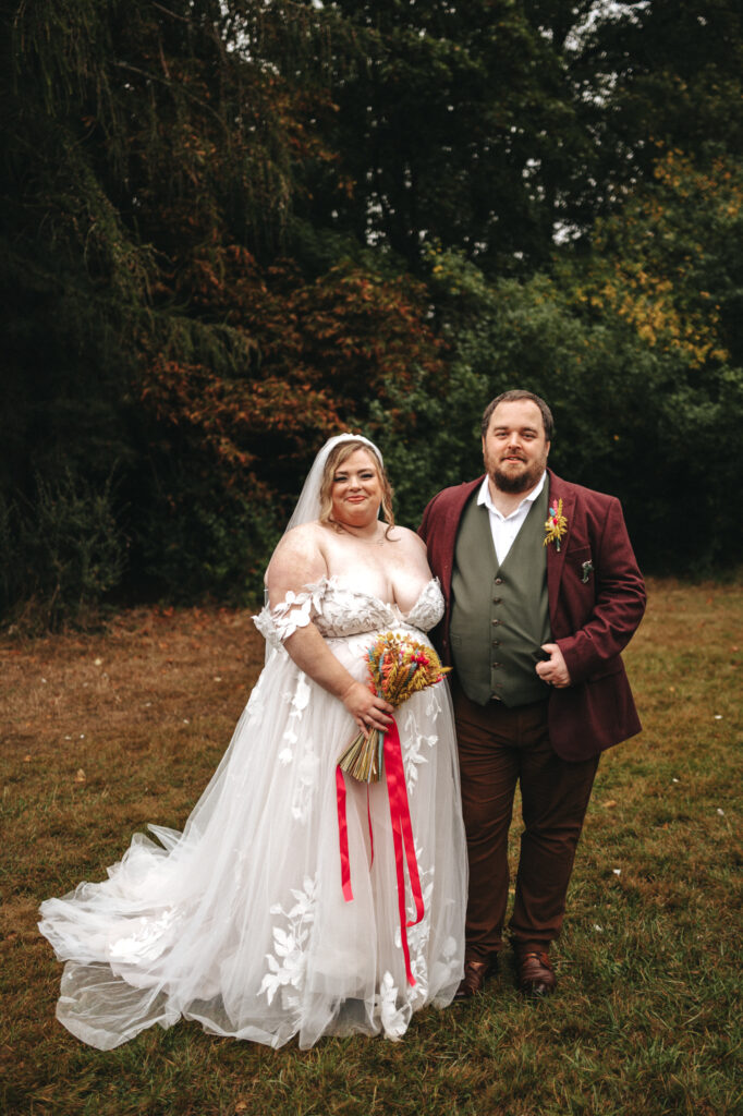 A bride and groom stand side by side outdoors, reminiscent of a Lord of the Rings scene. The bride wears a flowing white dress with floral details and off-the-shoulder sleeves, holding a bouquet with red ribbons. The groom dons a brown blazer over a green vest and white shirt, framed by greenery and majestic trees. © Aimee Lince Photography