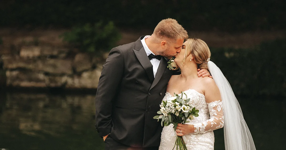 A couple stands by a tranquil body of water, captured by a skilled wedding photographer. The groom, in a black tuxedo, gently kisses the bride's forehead as she holds a bouquet of white flowers. Her off-shoulder gown and veil flutter softly against the lush greenery in the background. © Aimee Lince Photography - Wedding photographer in Lincolnshire, Yorkshire & Nottinghamshire