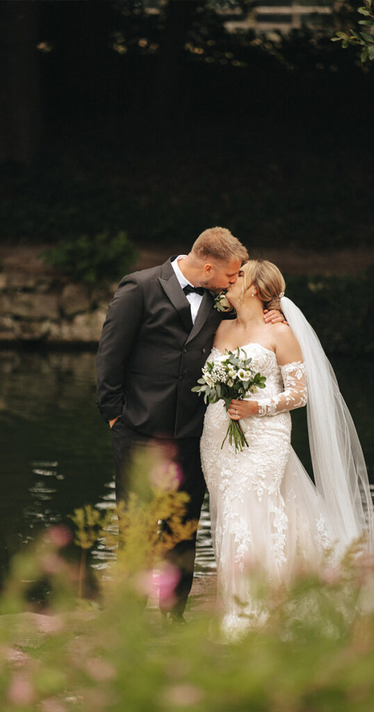 A bride and groom share a kiss beside a tranquil lake, lovingly captured by their wedding photographer. The bride wears an off-shoulder lace gown and veil, holding a bouquet of white flowers. The groom is in a dark suit with a bow tie. Greenery surrounds them, adding a natural backdrop to this romantic scene. © Aimee Lince Photography