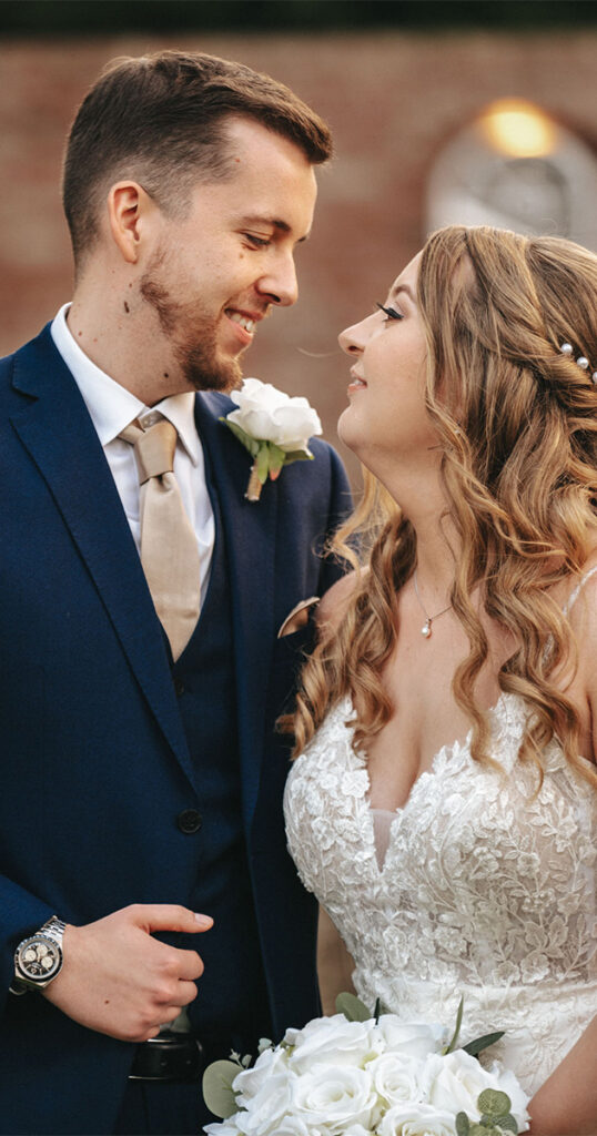 A bride and groom gaze at each other lovingly during their wedding. The groom wears a navy suit with a beige tie and white rose boutonniere. The bride is in a lace dress, wavy adorned hair, holding white roses. They stand outdoors by Stallingborough Grange, with a blurred brick wall and window behind them. © Aimee Lince Photography