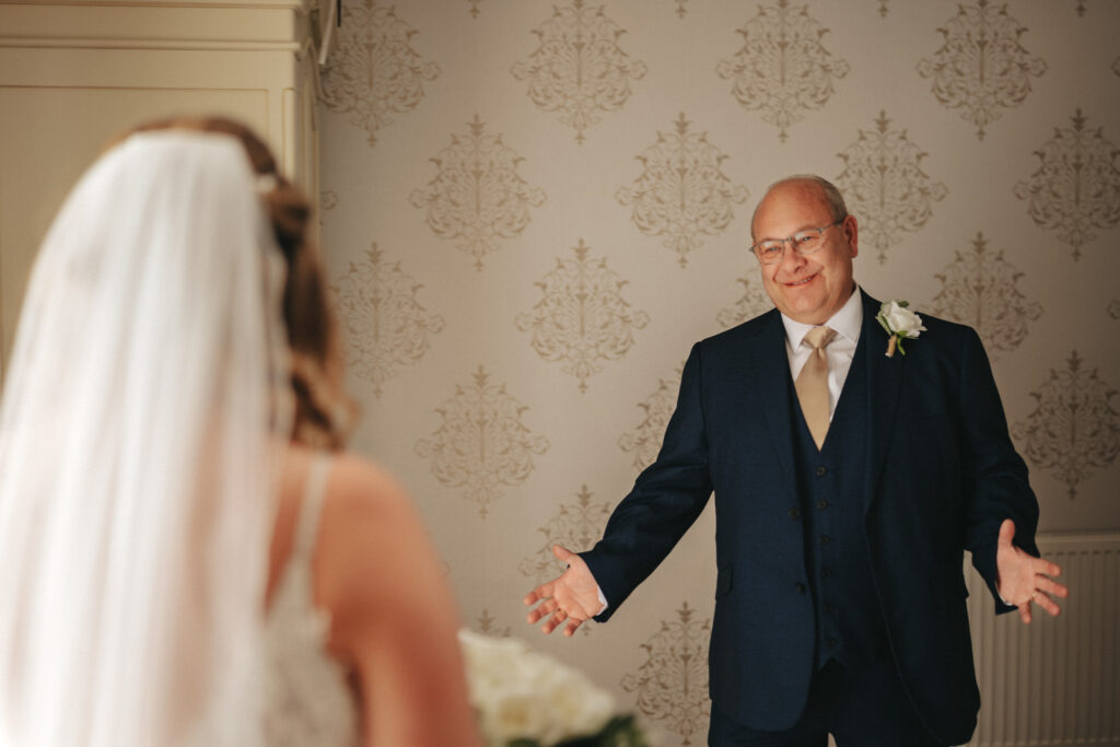 A man in a dark suit with a white shirt and gold tie smiles with open arms, a white rose boutonniere pinned on his lapel. In the foreground, the bride in her wedding dress and veil is slightly blurred against the elegant wallpaper, capturing a joyful moment at Stallingborough Grange. © Aimee Lince Photography