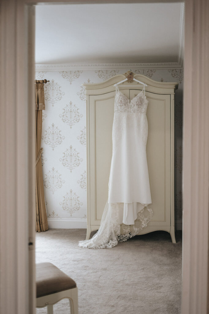 A white, lace wedding dress hangs on an ornate, cream-colored wardrobe door in an elegantly decorated room at Stallingborough Grange. The room features patterned wallpaper, beige carpet, and a gold curtain to the side. The photo is framed by a doorway, adding to the intimate, serene atmosphere. © Aimee Lince Photography