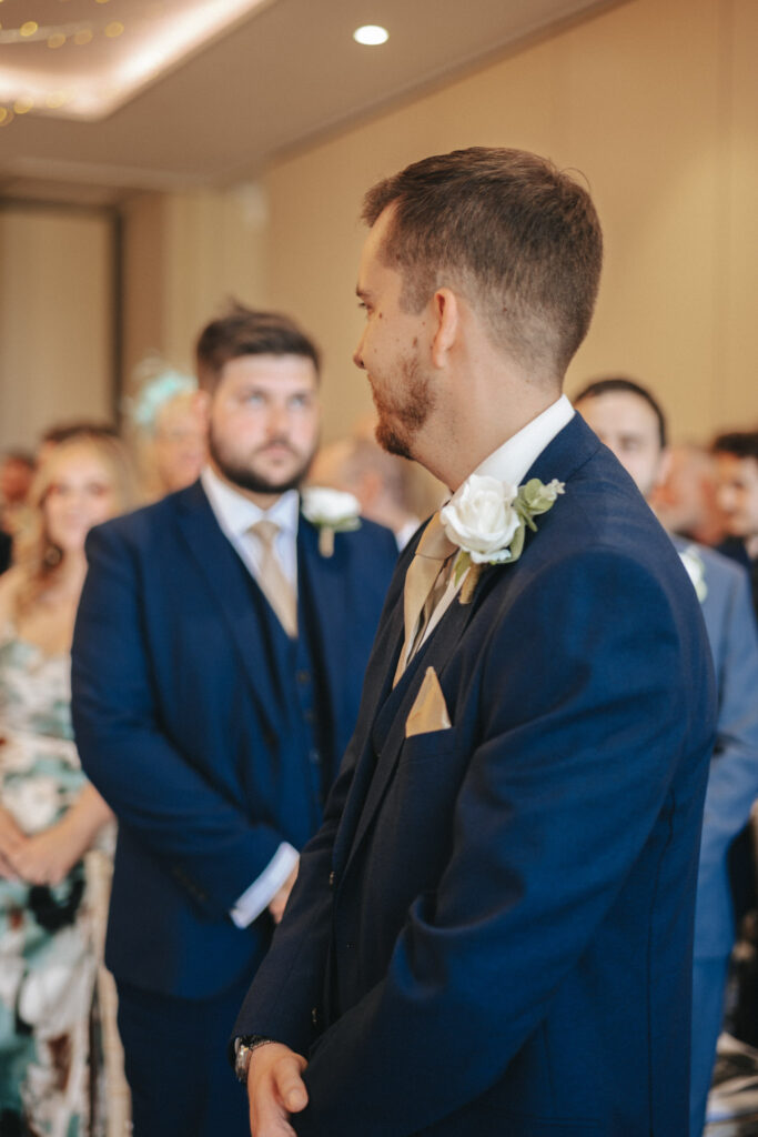 A groom in a navy suit with a white rose boutonniere looks forward during the wedding ceremony at Stallingborough Grange. Guests in formal attire, including a man in a matching suit and a woman in a floral dress, blur softly into the warmly lit elegance of the room. © Aimee Lince Photography - Wedding photographer in Lincolnshire, Yorkshire & Nottinghamshire