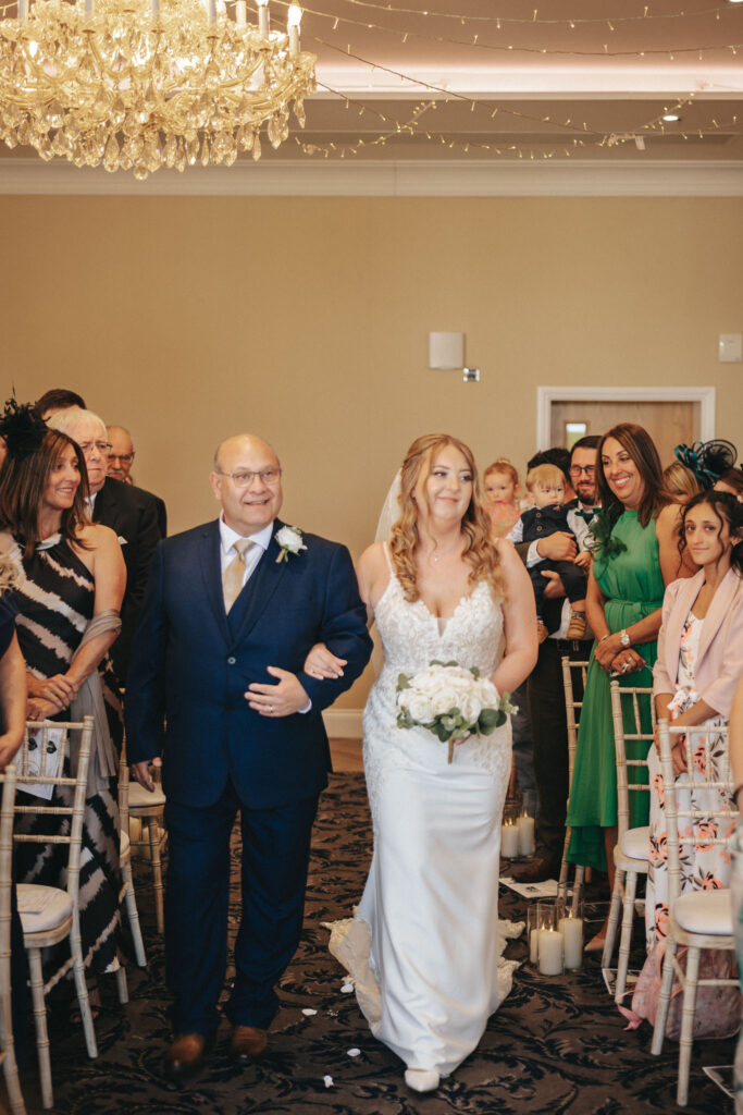 At the Stallingborough Grange, a radiant bride in a white gown holding a bouquet walks down the aisle with a man in a blue suit. Guests seated on white chairs gaze upon them as candles and fairy lights add to the ambiance beneath an elegant chandelier amidst scattered petals. © Aimee Lince Photography - Wedding photographer in Lincolnshire, Yorkshire & Nottinghamshire