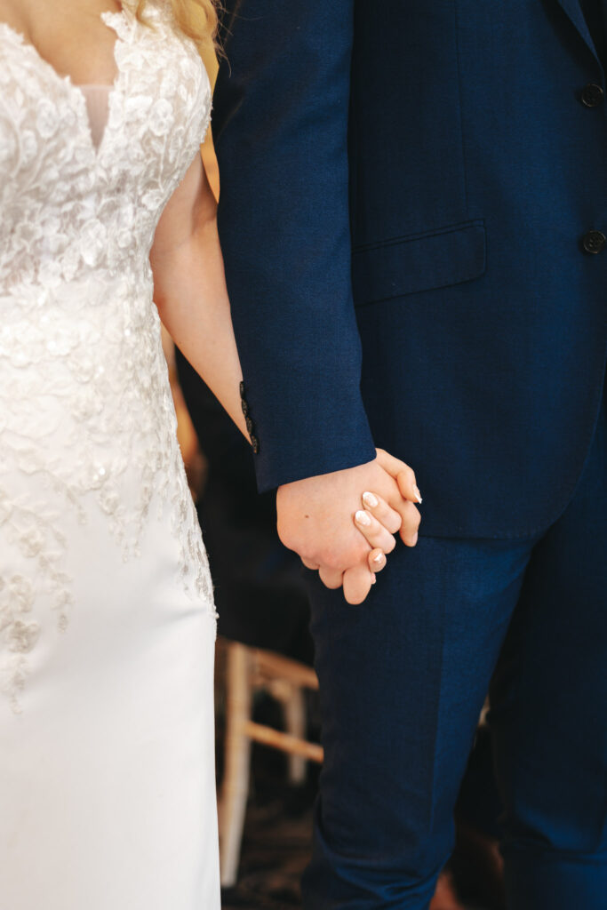 At Stallingborough Grange, a couple holds hands during a wedding ceremony. The bride is radiant in her white lace dress, and the groom looks sharp in his navy blue suit. Their hands are joined, embodying a moment of togetherness and commitment amid the softly blurred background. © Aimee Lince Photography - Wedding photographer in Lincolnshire, Yorkshire & Nottinghamshire