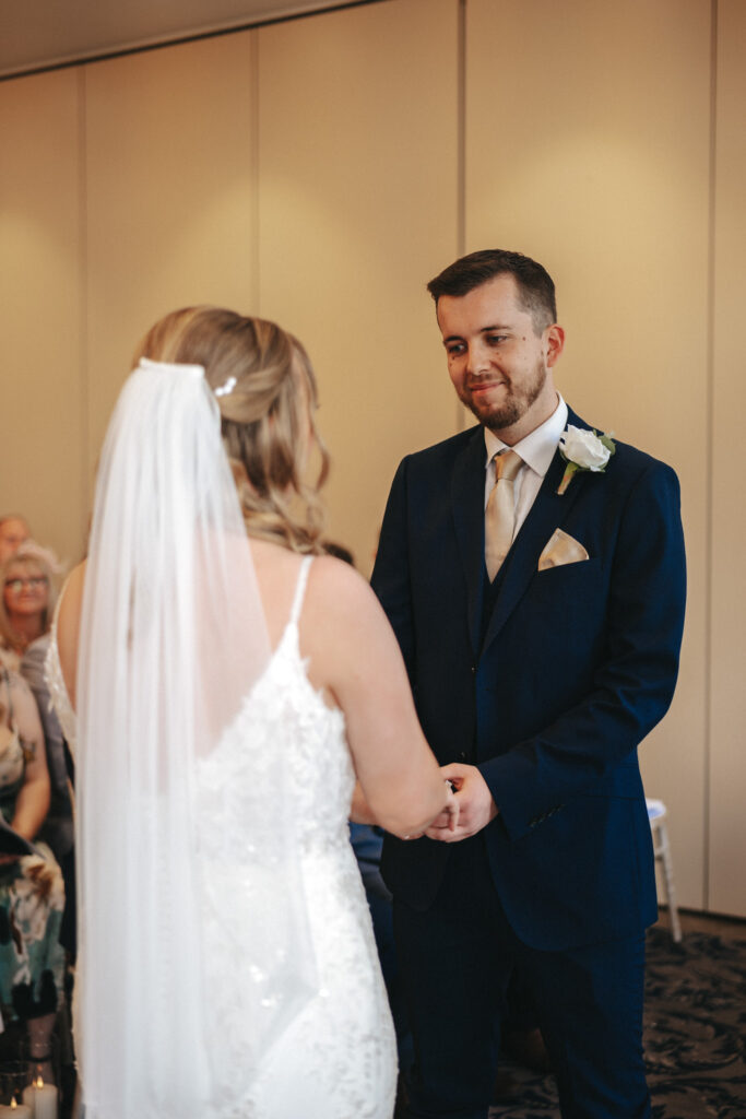 A couple stands facing each other during their Stallingborough Grange wedding. The bride, in a long veil and lace dress, holds the groom's hands. He wears a dark blue suit with a tan tie and white rose boutonniere. Guests are seated in soft focus in the background. © Aimee Lince Photography - Wedding photographer in Lincolnshire, Yorkshire & Nottinghamshire