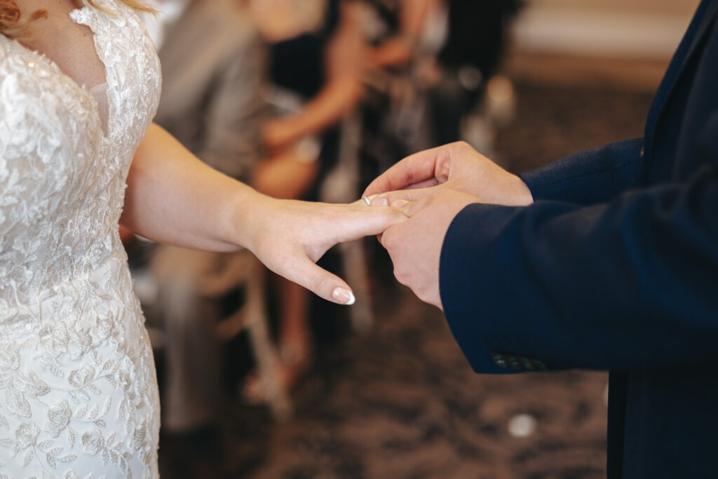 Close-up of a wedding ceremony at Stallingborough Grange, where a bride in a lace white dress is having a ring placed on her finger by another person in a navy suit. The background shows blurred guests seated, watching the exchange of vows and rings. © Aimee Lince Photography - Wedding photographer in Lincolnshire, Yorkshire & Nottinghamshire