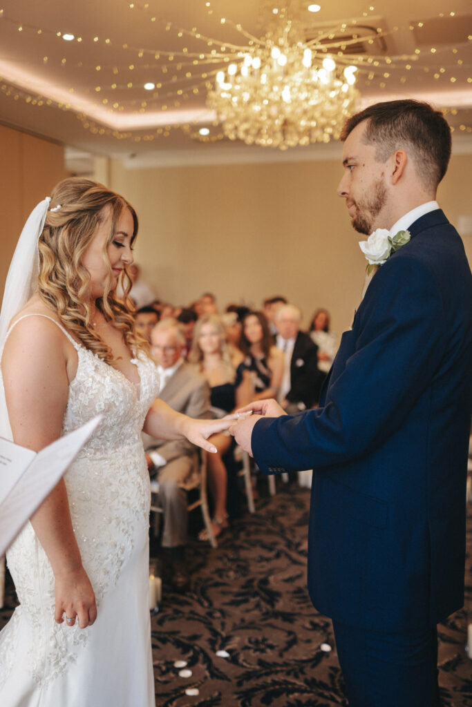 At Stallingborough Grange, a bride and groom exchange rings during their wedding ceremony. The bride wears a white dress with lace details and a veil, while the groom is in a blue suit with a white rose boutonniere. They stand under a chandelier, with guests seated in the background. © Aimee Lince Photography - Wedding photographer in Lincolnshire, Yorkshire & Nottinghamshire