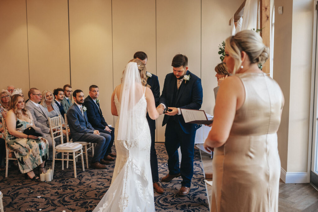 A bride in a white gown stands next to a groom in a dark suit during the wedding ceremony at Stallingborough Grange. They are surrounded by guests seated in rows, while an officiant, holding a book, prepares to conduct the ceremony. The room features beige walls and a dark floral-patterned carpet. © Aimee Lince Photography - Wedding photographer in Lincolnshire, Yorkshire & Nottinghamshire