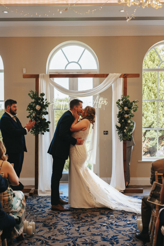 A bride and groom share a kiss at their wedding ceremony beneath a wooden arch adorned with white fabric and greenery. Behind them, a neon sign reads "Happily Ever After." Set indoors at Stallingborough Grange, large arched windows frame the scene as guests watch from both sides. © Aimee Lince Photography - Wedding photographer in Lincolnshire, Yorkshire & Nottinghamshire