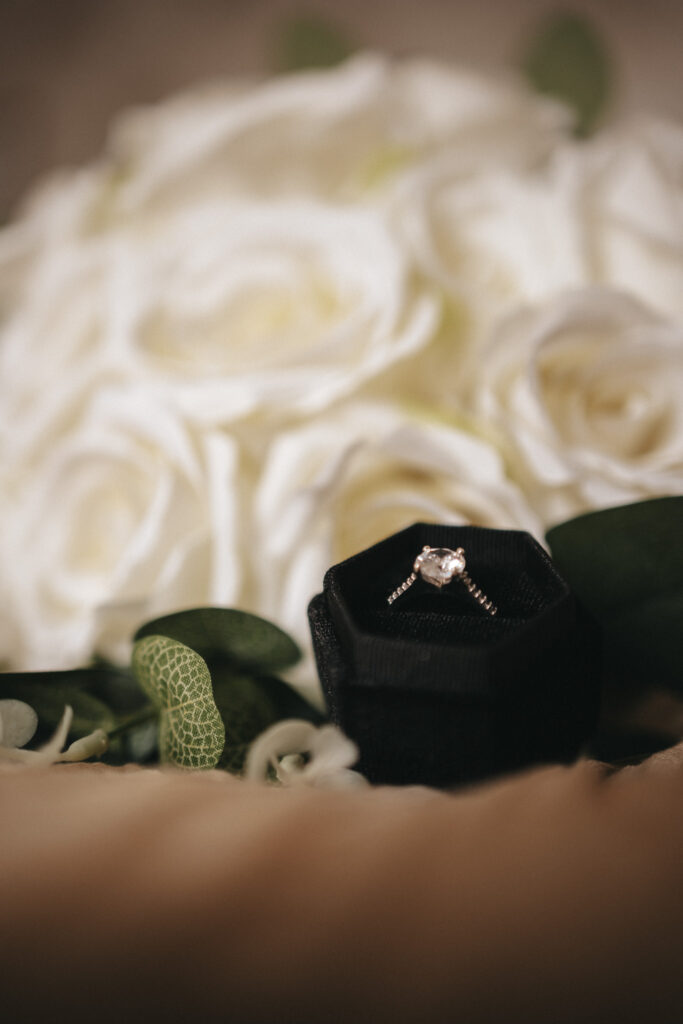 A diamond engagement ring in a black hexagonal velvet box is in the foreground. The background features a blurred bouquet of white roses with green leaves, creating a soft and romantic setting—perfect for any bride about to celebrate her special day at Stallingborough Grange. © Aimee Lince Photography